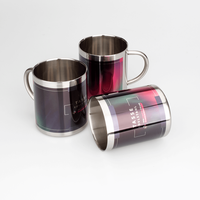 drei Edelstahltassen, vordere liegend mit Fotodruck, zwei anderen stehen seitlich und man sieht ihren Henkel